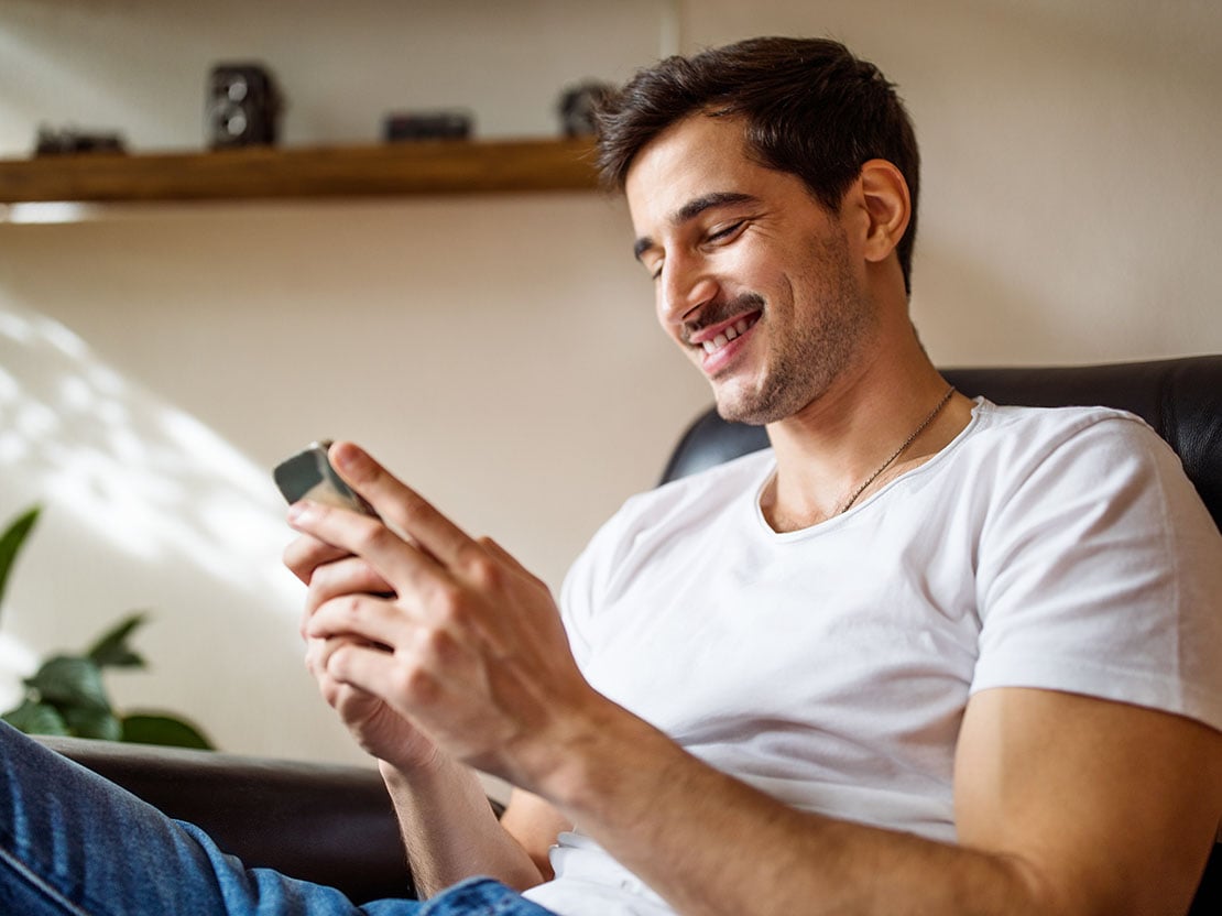 Man i vit t-shirt tittar ned i sin mobil och ler